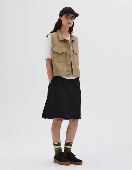 Full body image, front view of model wearing sleeveless beige shirt with corozo button closure and flap chest pockets. Layered over a white t-shirt and styled with black skirt and cap, with green striped socks and black nubuck trainers.
