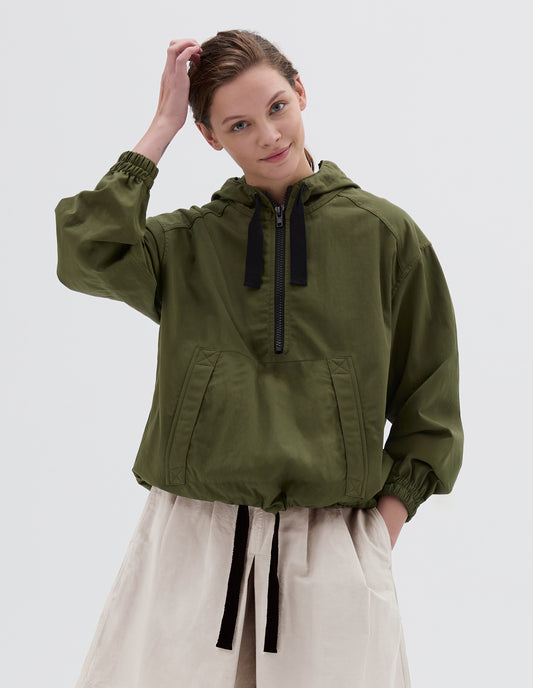 Cropped view of model wearing a green drawcord hooded jacket, with kangaroo pocket and half zip fastening. Styled with beige shorts.
