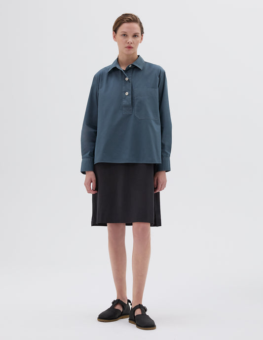 Full body image, front view of model wearing long sleeve blue shirt with half placket, corozo buttons and large chest pocket. Styled with black skirt and black leather t-bar sandals. 