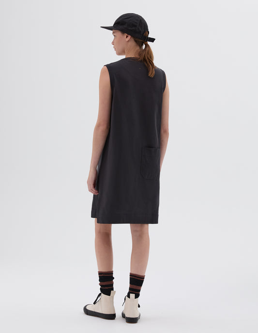 Full body image, back view of model wearing black regular cut knee length sleeveless and collarless dress with split side hem and patch back pocket, styled with black cap, black socks with white and black high top trainers.