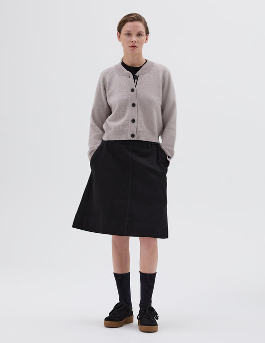 Full body image, front view of model wearing grey cropped length cardigan with raglan sleeve, black corozo brace button fastening, contrast stripe on inside placket, styled with black t-shirt and dark blue skirt with side pockets and black socks with black nubuck trainers.