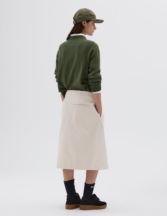 Full body image, back view of model wearing white midi length skirt with welt back pocket and side pockets, styled with green cap, white collared shirt, green knitted jumper and black socks with black nubuck trainers.