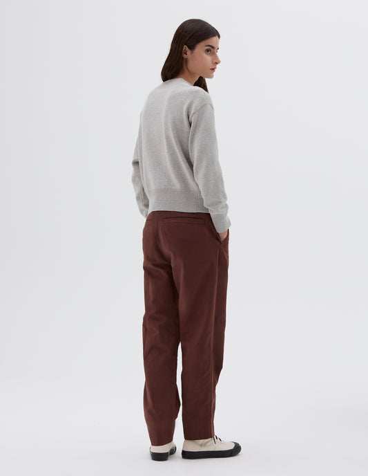 Full body image, back view of model wearing brown ankle length tapered trousers with side pockets and welt back pockets, styled with light grey knitted jumper, black leather belt and black and white trainers. 