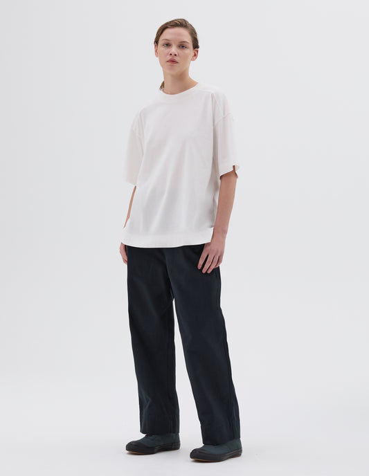 Full body image, front view of model wearing white relaxed long t-shirt with side split hem, styled with dark blue trousers and dark grey trainers.
