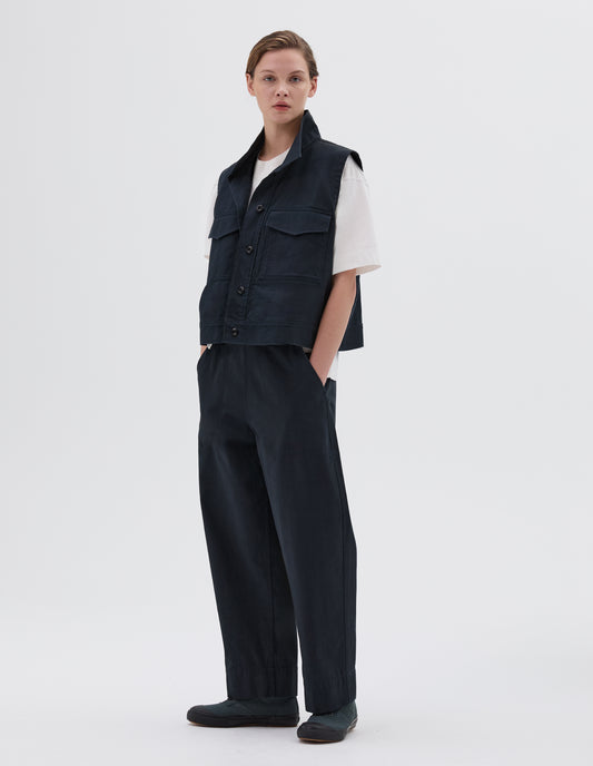 Full body image, front view of model wearing sleeveless navy shirt with corozo button closure and flap chest pockets. Layered over a white t-shirt and styled with navy trousers and green canvas trainers.