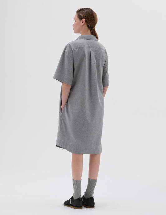 Full body image, back view of model wearing grey below the knee shirt dress with short sleeves, side pockets and back pleat, styled with light grey socks and black leather sandals.