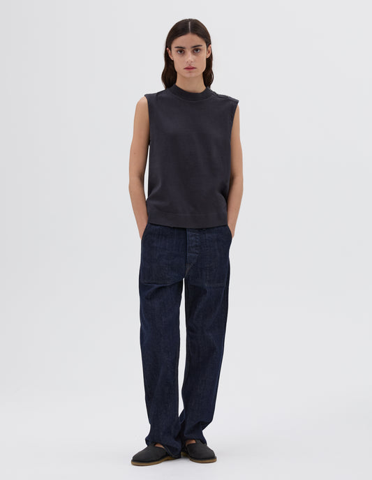 Full body image, front view of model wearing dark blue regular cut sleeveless top with button-through shoulder with corozo brace buttons and ribbed neckline, styled with blue jeans and black leather sandals.