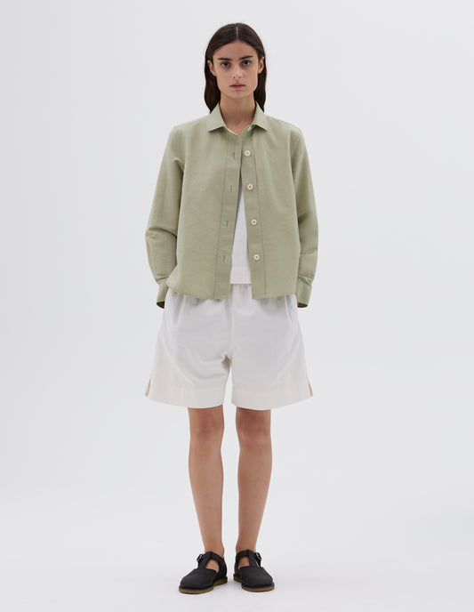 Full body image, front view of model wearing green long sleeve shirt with corozo buttons. Layered over a white vest and worn with white shorts and black leather t-bar sandals.