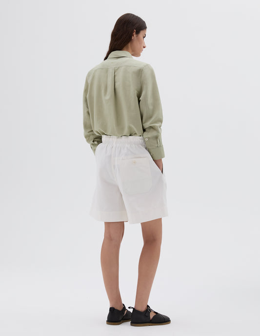 Full body image, back view of model wearing white relaxed cut shorts with elasticated waist, side pockets, button-through patch pocket, styled with light green long-sleeved shirt and black leather sandals.
