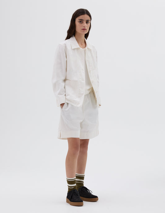 Full body image, front view of model wearing off white jacket with two patch pockets, button closure and cuffs. Styled over white vest, with white shorts, striped socks and black nubuck trainers. 