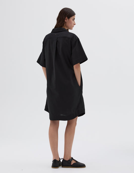 Full body image, back view of model wearing black below the knee shirt dress with short sleeves, side pockets and back pleat, styled with black leather sandals.
