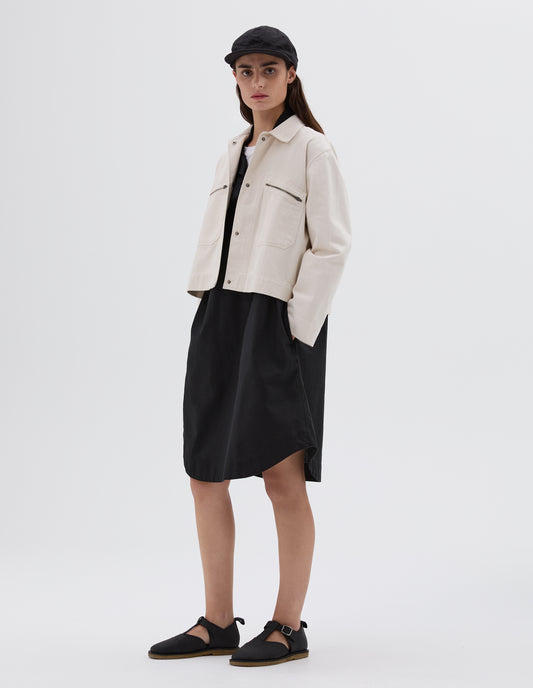 Full body image, front view of model wearing white cropped jacket with two front zip pockets and concealed press stud fastening. Styled over a black v-neck dress and white t-shirt, with black cap and black leather t-bar sandals. 