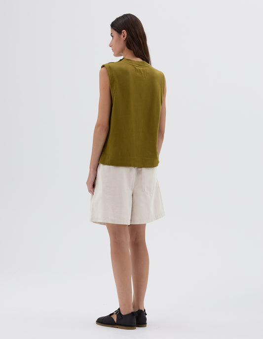 Full body image, back view of model wearing green regular cut sleeveless top with ribbed neckline and side split hem, styled with white pleated shorts and black leather sandals.