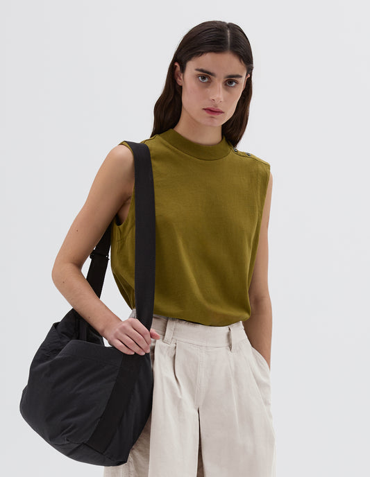 Cropped body image, front view of female model wearing navy blue bag with contrast black herringbone tape shoulder strap, large popper fastening pocket and Velcro closure. Styled with olive green sleeveless vest and beige shorts.