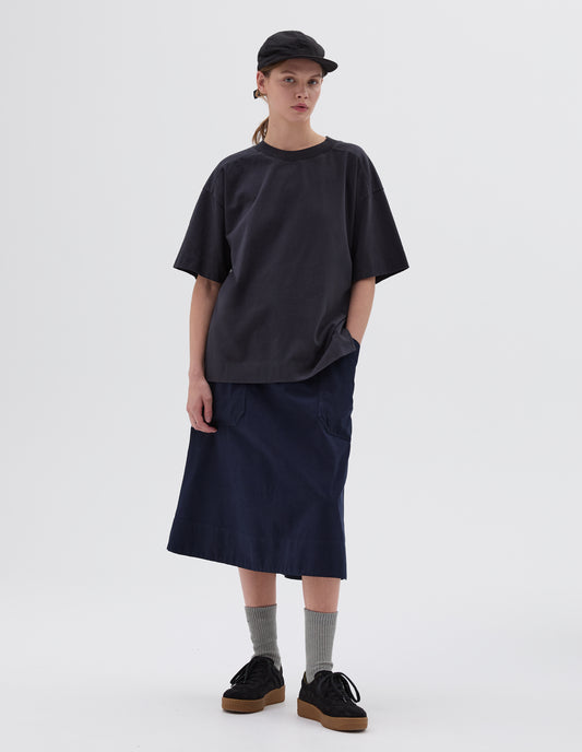 Full body image, front view of model wearing dark blue relaxed long t-shirt with side split hem, styled with black cap and dark blue skirt with large patch pockets, light green socks and black nubuck trainers. 