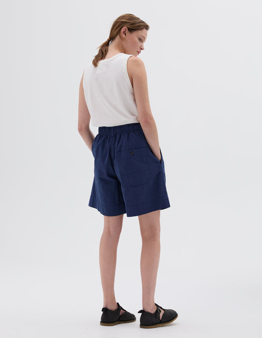 Full body image, back view of model wearing blue relaxed cut shorts with elasticated waist, side pockets, button-through patch pocket, styled with white sleeveless top and black leather sandals. 
