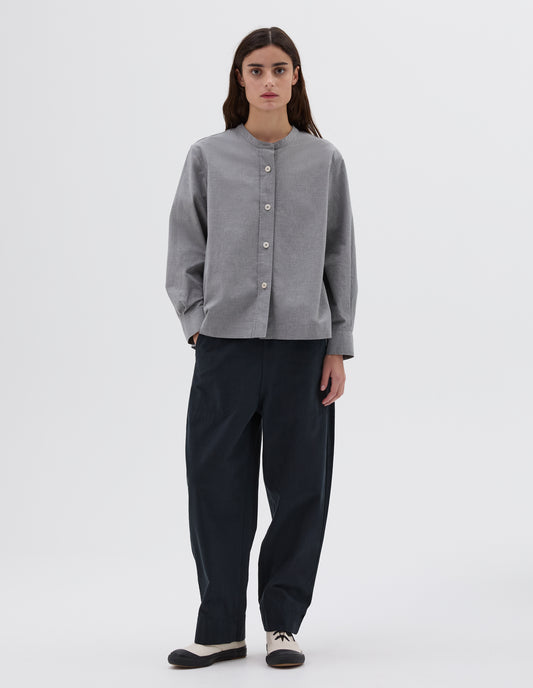 Full body image, front view of model wearing collarless long sleeve grey shirt with corozo button closure. Styled with navy trousers and white canvas trainers. 