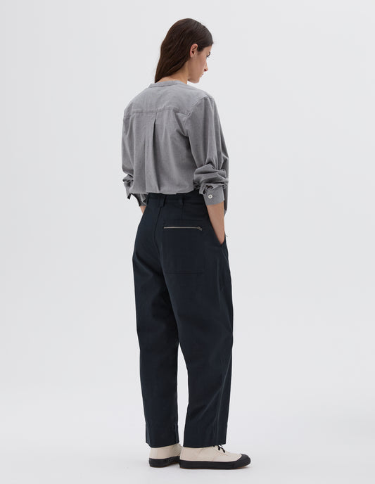 Full body image, back view of model wearing dark blue straight cut ankle crop trousers with side pockets, zip patch back pocket and belt loops, styled with light grey long sleeved collarless shirt and white and black trainers.
