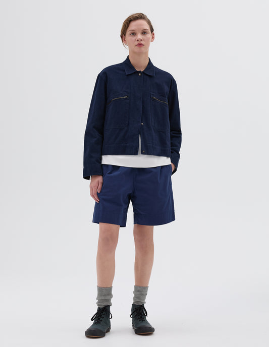 Full body image, front view of model wearing indigo blue cropped jacket with two front zip pockets and concealed press stud fastening. Styled over a white vest, with blue shorts, grey socks and green high top trainers.