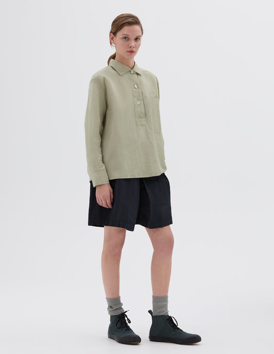 Full body image, front view of model wearing long sleeve light green shirt with half placket, corozo buttons and large chest pocket. Styled with navy pleated shorts, grey socks and green high top trainers.