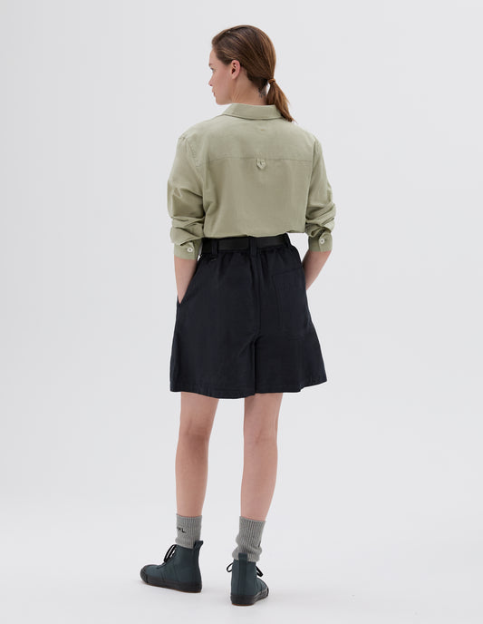 Full body image, back view of model wearing dark grey relaxed shorts with three belt loops with one D-ring detail, side pockets and back pocket, styled with leather belt, light green long-sleeved collared shirt, grey socks and dark grey high top trainers.