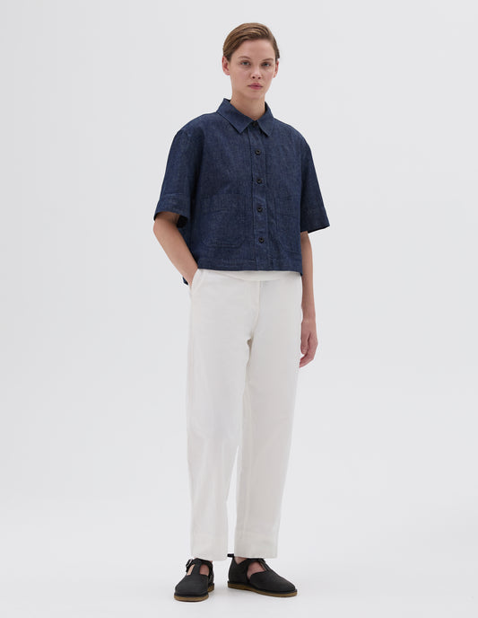 Full body image, front view of model wearing cropped indigo short sleeve shirt with corozo button closure and two patch pockets. Styled with white trousers and black leather t-bar sandals. 
