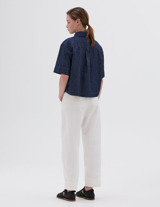Full body image, back view of model wearing cropped indigo short sleeve shirt with hanger loop on back yoke. Styled with white trousers and black leather t-bar sandals. 