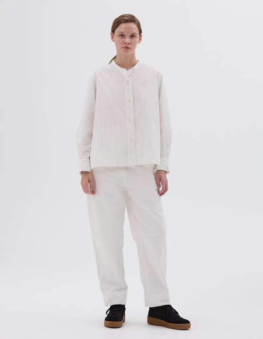Full body image, front view of model wearing collarless long sleeve white and olive green striped shirt with corozo button closure. Styled with white trousers and black nubuck leather trainers. 