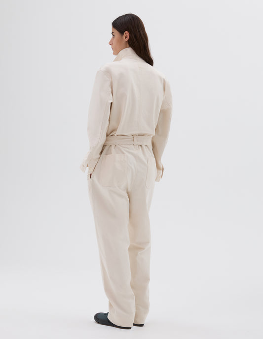 Full body image, back view of model wearing relaxed white overalls with   belt and belt loops, patch back pockets and corozo brace buttons on cuffs, and side pockets. Styled with dark grey trainers.