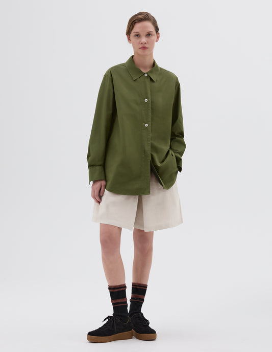 Full body image, front view of model wearing long sleeve, longline green shirt with corozo button closure and cuffs. Styled with beige pleated shorts, brown and black socks and black nubuck leather trainers. 