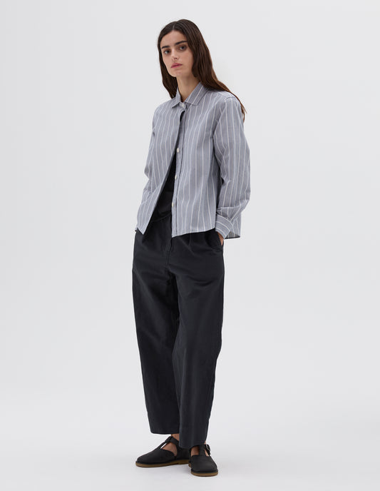 Full body image, front view of model wearing blue and white striped long sleeve shirt with corozo buttons. Layered over a navy t-shirt and styled with navy trousers and black leather t-bar sandals. 