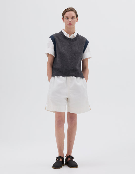 Full body image, front view of model wearing grey knitted slipover with ribbed hem and neckline and blue ribbed armholes, styled with white short sleeved collared shirt and white shorts and black leather sandals.