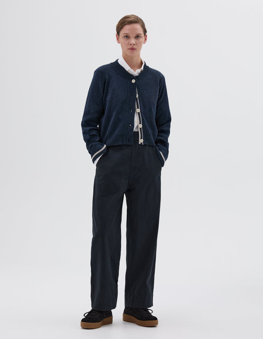 Full body image, front view of model wearing blue cropped length cardigan with raglan sleeve, white corozo brace button fastening, contrast stripe on inside placket and on cuffs, styled with white shirt and dark blue trousers with side pockets and black nubuck trainers.
