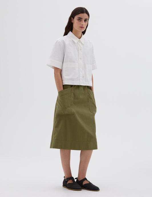 Full body image, front view of model wearing cropped white short sleeve shirt with corozo button closure and two patch pockets. Styled with green patch pocket skirt and black leather t-bar sandals. 