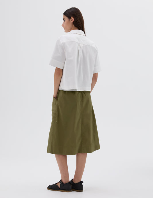 Full body image, front view of model wearing cropped white short sleeve shirt with box pleat and hanger loop on back yoke. Styled with green patch pocket skirt and black leather t-bar sandals. 