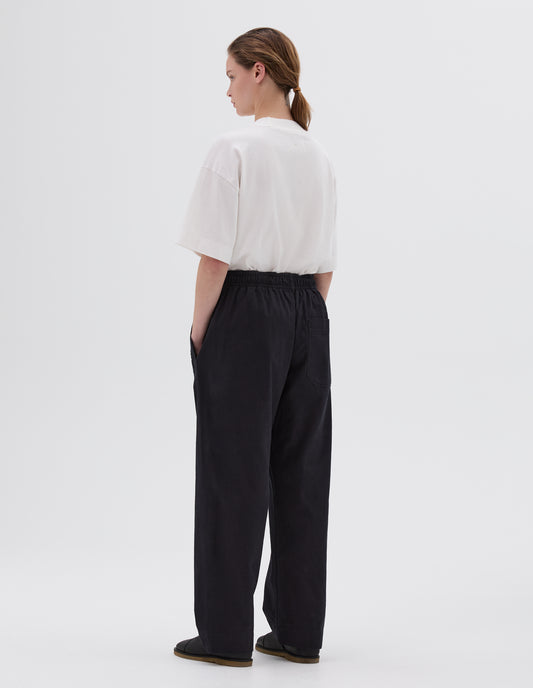 Full body image, back view of model wearing relaxed dark blue joggers with elasticated waist and patch back pocket. Styled with white t-shirt and black leather sandals.