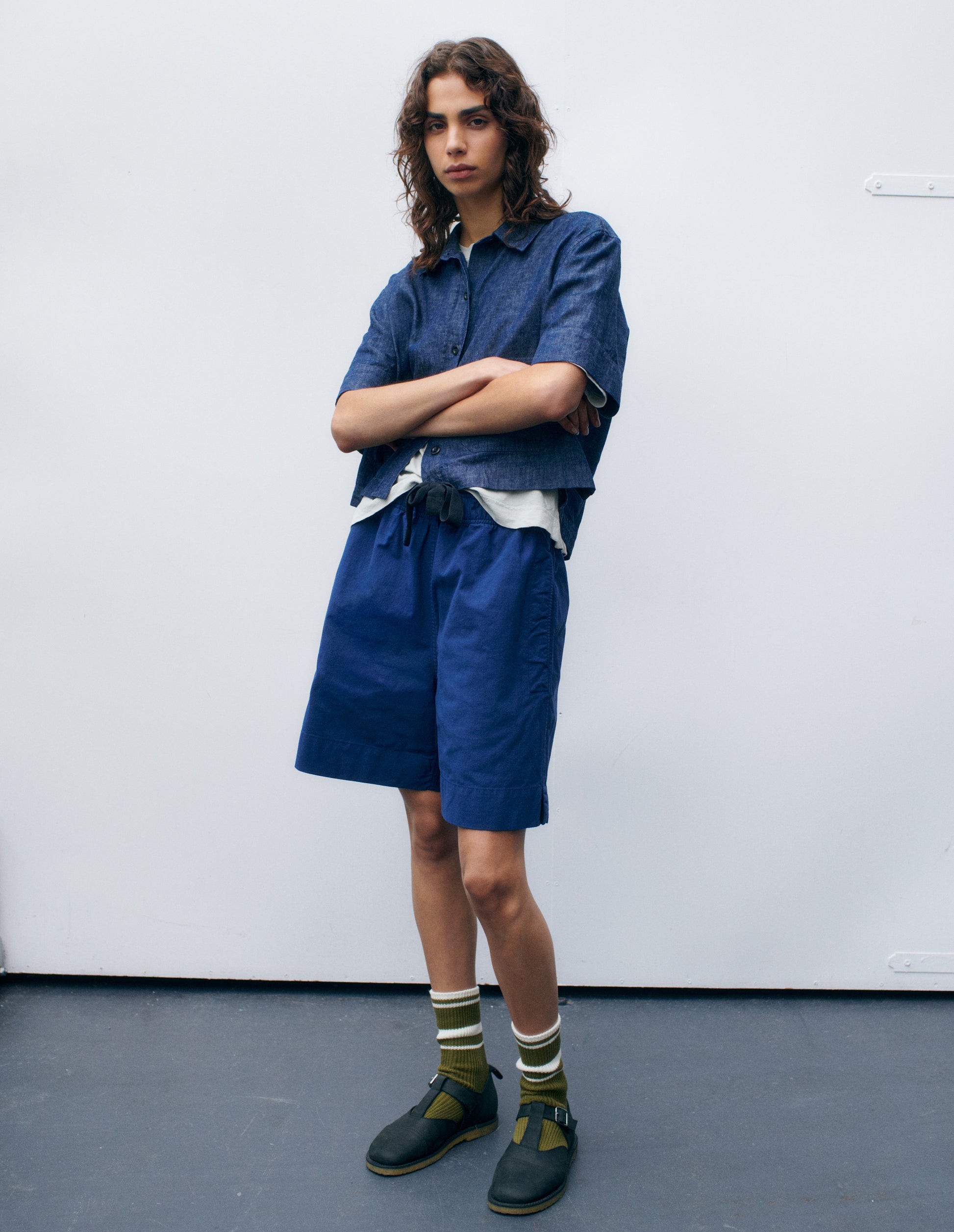 Full body image, front view of model wearing indigo blue short sleeve shirt, layered over white t-shirt. Styled with blue shorts, green striped socks and black leather t-bar sandals.