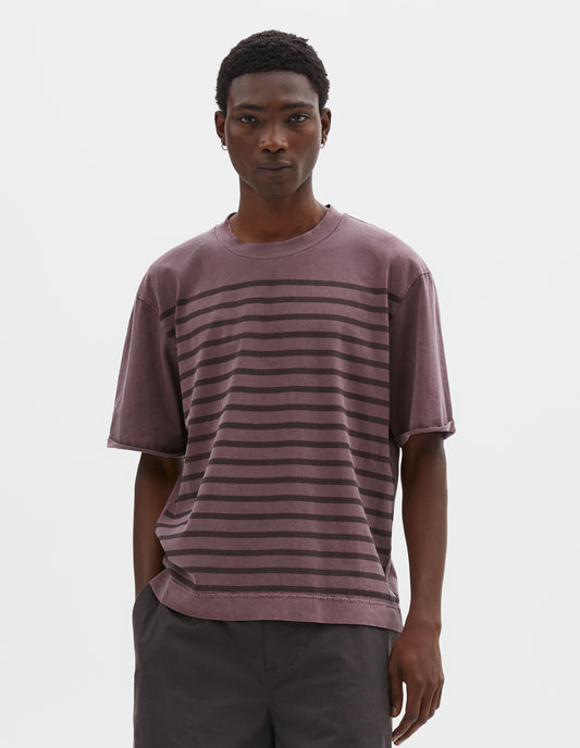 Cropped body image, front view of model wearing purple short sleeve cotton t-shirt with brown stripes, flatlock stitch seam detailing and rolled raw sleeve hems held with a single bar tack. Styled with dark brown shorts.