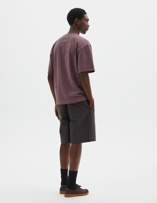 Full body image, back view of model wearing purpleish pink short sleeve t shirt with flatlock stitch seam detailing and rolled raw hems held with a single bar tack. Styled with brown shorts, black socks and brown suede trainers and gum soles. 
