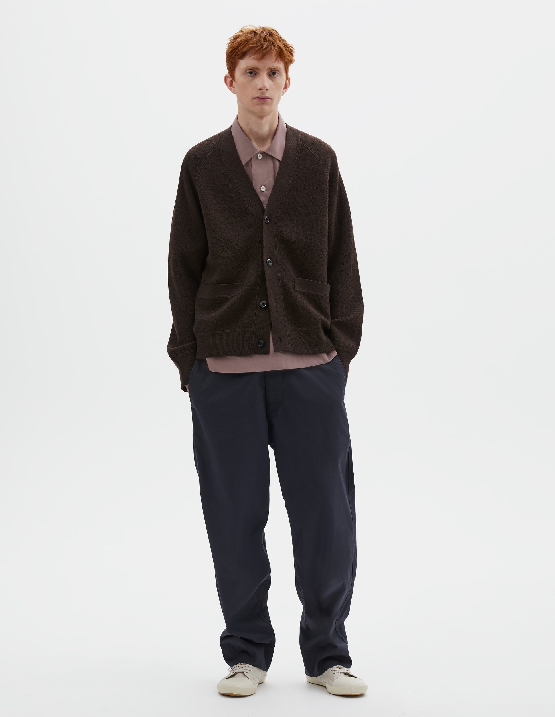 Full body image, front view of model wearing brown v neck wool cardigan, with concealed pockets, corozo button closure, raglan sleeves and contrast rib on cuffs and hem. Styled over dark pink shirt, dark blue trousers and beige plimsolls.