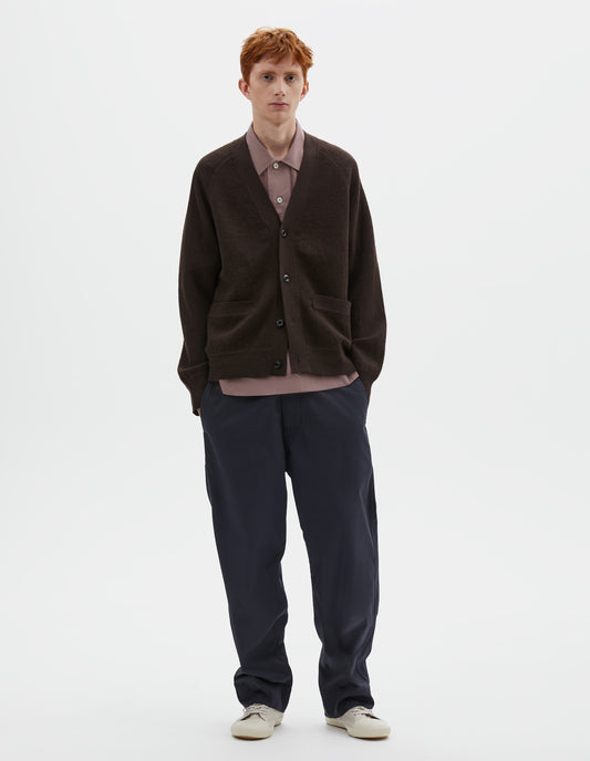Full body image, front view of model wearing brown v neck wool cardigan, with concealed pockets, corozo button closure, raglan sleeves and contrast rib on cuffs and hem. Styled over dark pink shirt, dark blue trousers and beige plimsolls.
