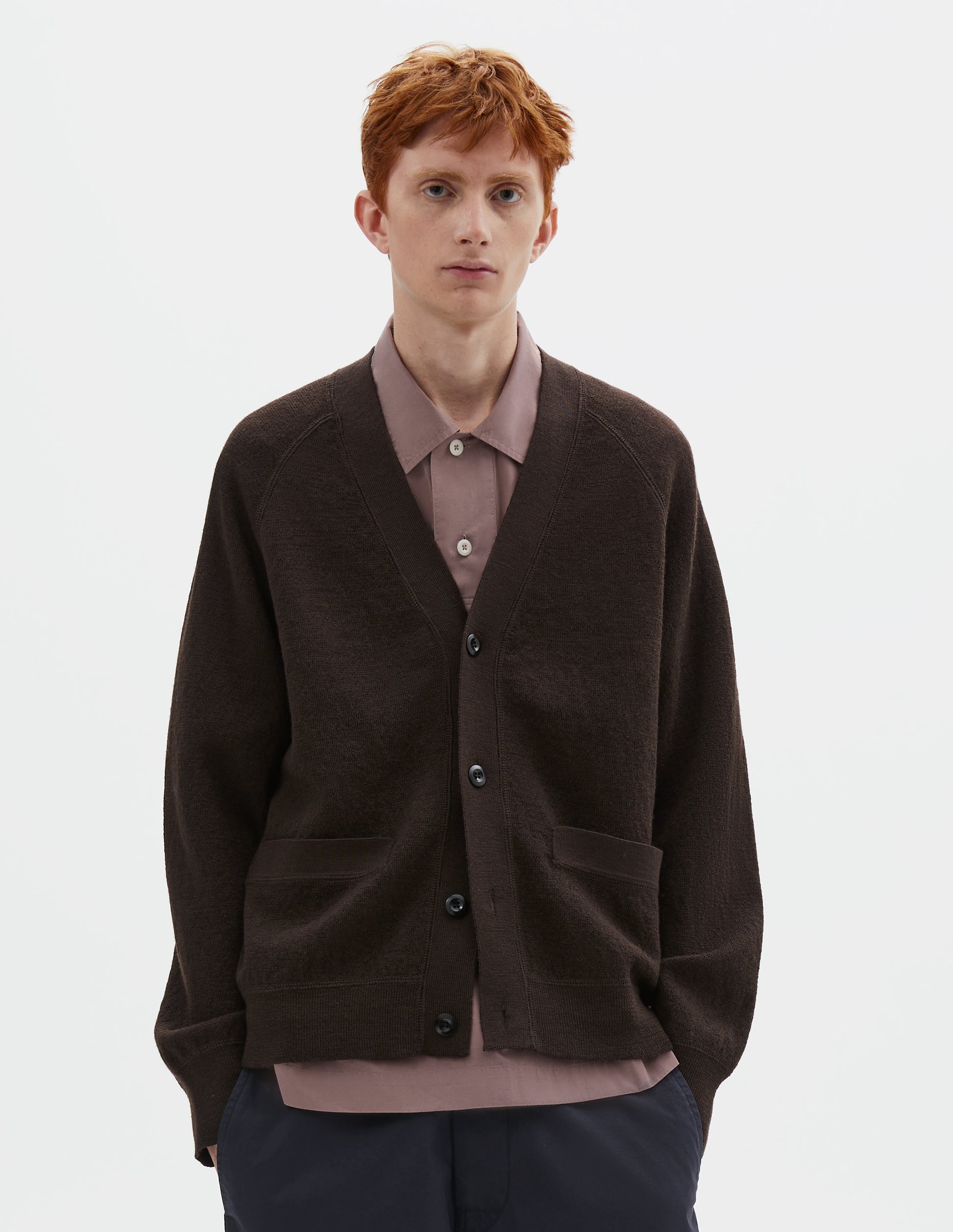 Cropped body image, front view of model wearing brown v neck wool cardigan, with concealed pockets, corozo button closure, raglan sleeves and contrast rib on cuffs and hem. Styled over dark pink shirt and dark blue trousers.