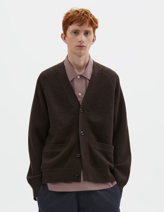 Cropped body image, front view of model wearing brown v neck wool cardigan, with concealed pockets, corozo button closure, raglan sleeves and contrast rib on cuffs and hem. Styled over dark pink shirt and dark blue trousers.