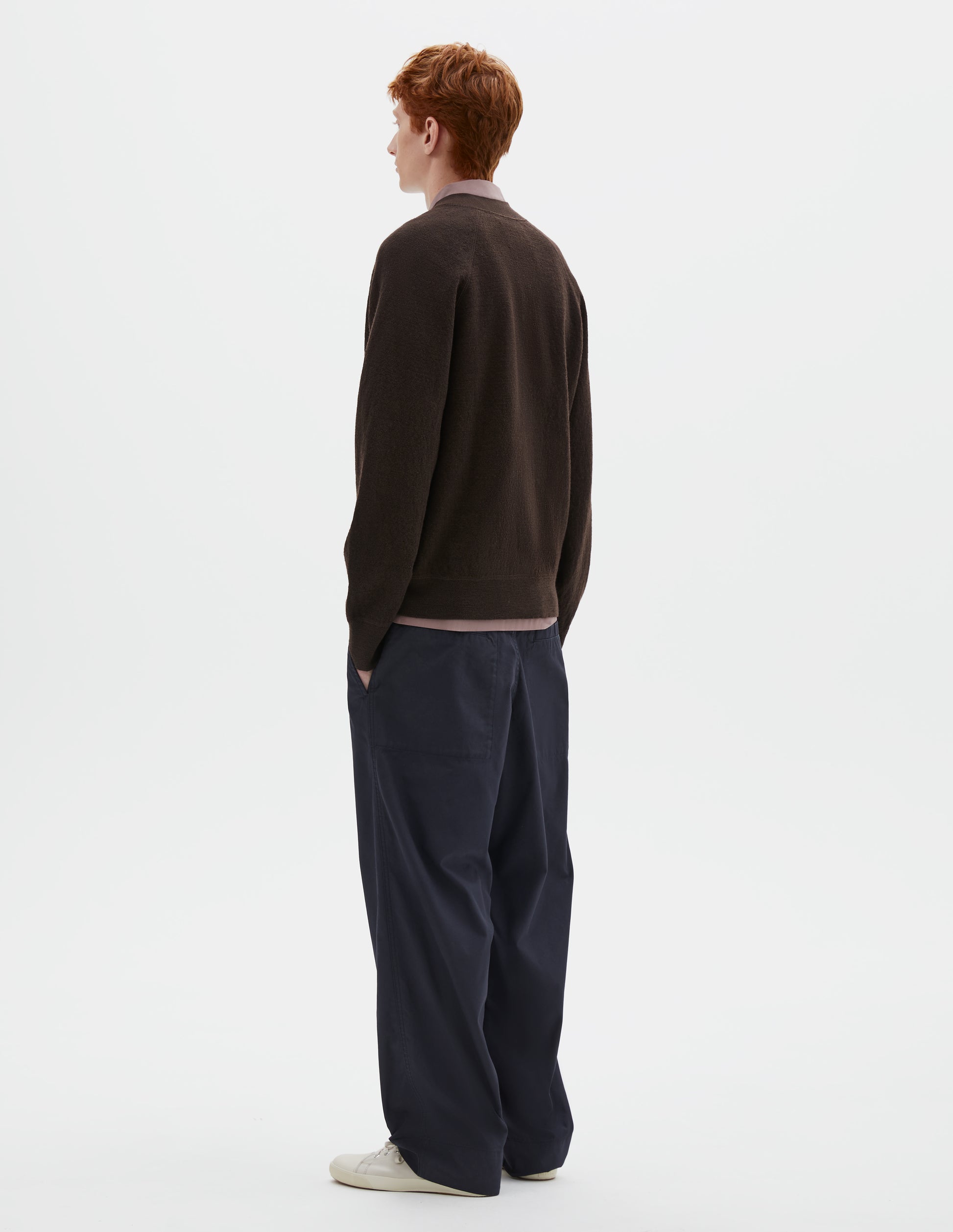 Full body image, back view of model wearing brown v neck wool cardigan, with raglan sleeves and contrast rib on cuffs and hem. Styled over dark pink shirt, dark blue trousers and beige plimsolls.