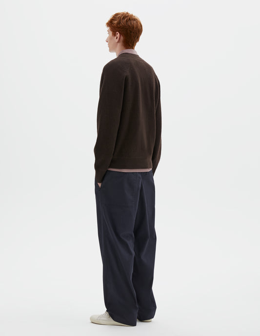 Full body image, back view of model wearing brown v neck wool cardigan, with raglan sleeves and contrast rib on cuffs and hem. Styled over dark pink shirt, dark blue trousers and beige plimsolls.