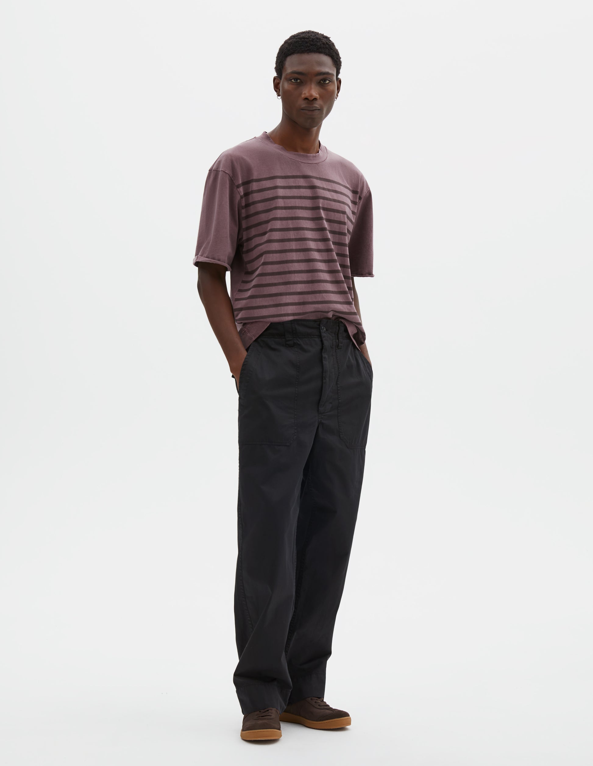 Full body image, front view of model wearing relaxed straight leg black trousers with sewn-in patch pockets, half-elasticated waistband with belt loops and corozo braze button fastening. Styled with dark lilac striped t-shirt and dark brown nubuck trainers.