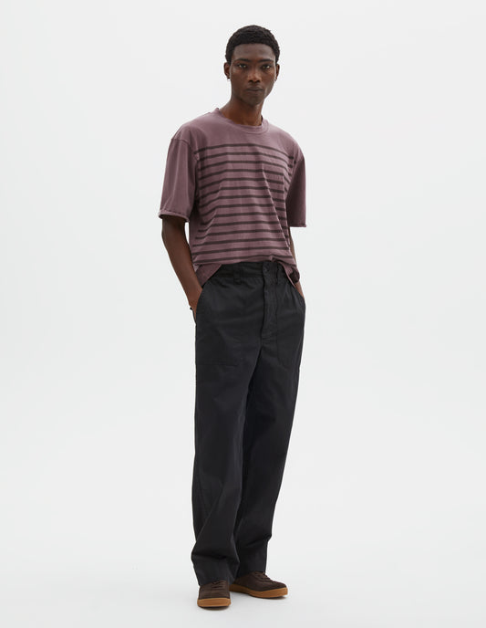 Full body image, front view of model wearing relaxed straight leg black trousers with sewn-in patch pockets, half-elasticated waistband with belt loops and corozo braze button fastening. Styled with dark lilac striped t-shirt and dark brown nubuck trainers.