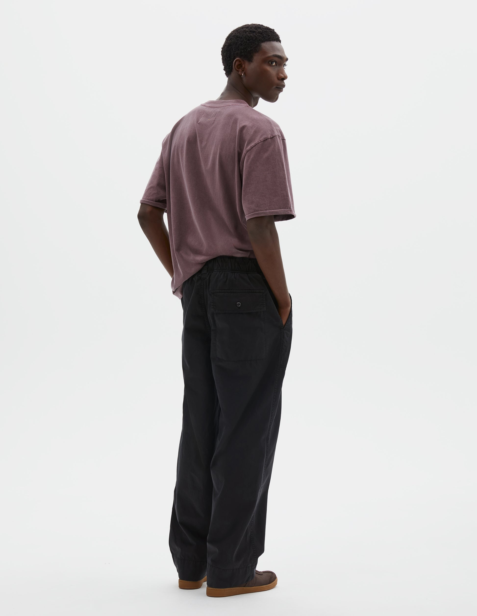 Full body image, back view of model wearing relaxed straight leg black trousers  with half-elasticated waistband, belt loops and corozo brace button fastening flap back pocket. Styled with dark lilac t-shirt and dark brown nubuck trainers.