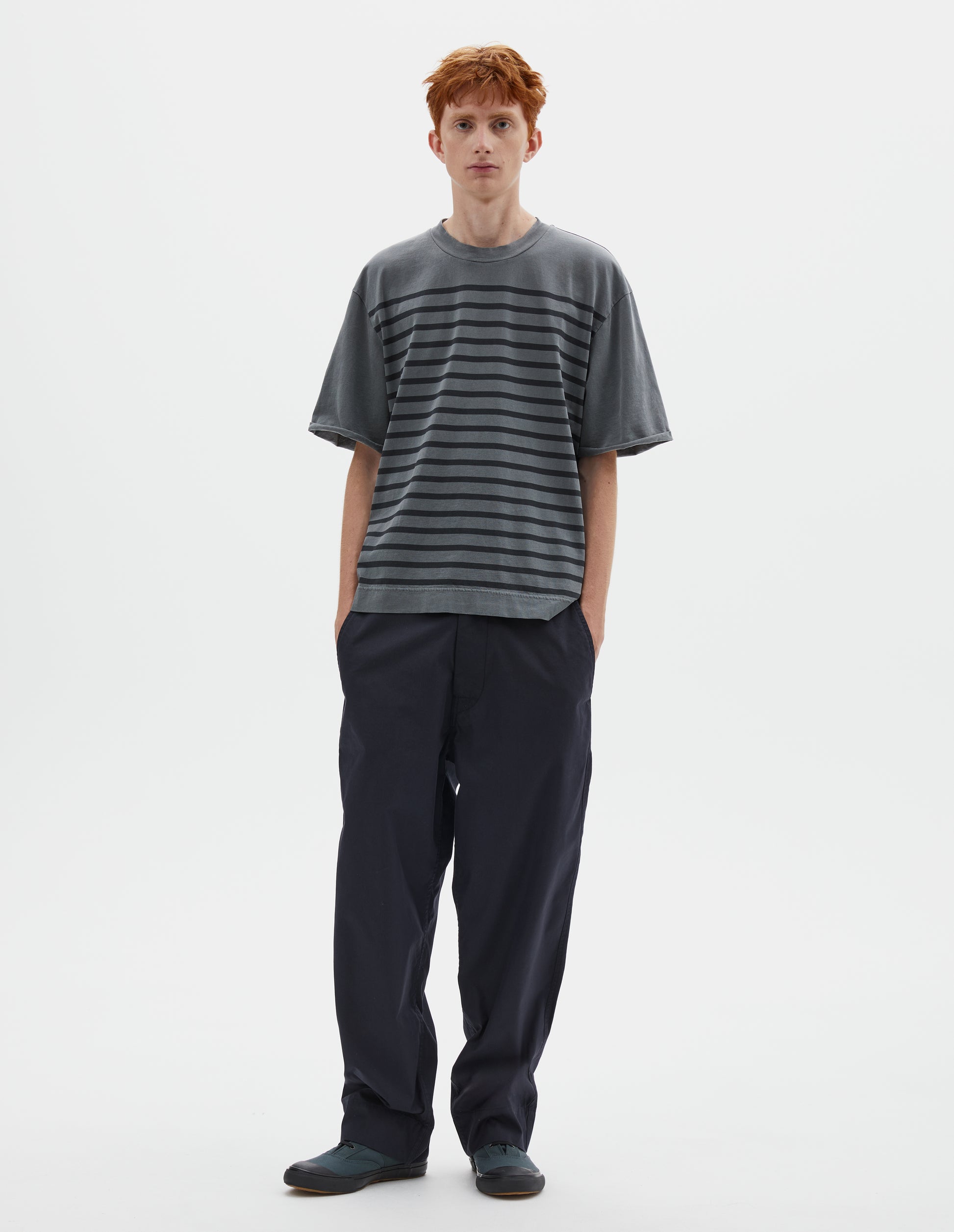 Full body image, front view image of model wearing blueish grey short sleeve t shirt with black stripes, flatlock stitch seam detailing and rolled raw sleeve hems held with a single bar tack. Styled with dark navy blue trousers and blue high top canvas trainers.  
