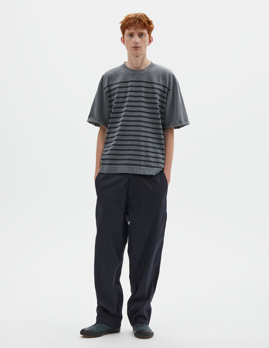 Full body image, front view image of model wearing blueish grey short sleeve t shirt with black stripes, flatlock stitch seam detailing and rolled raw sleeve hems held with a single bar tack. Styled with dark navy blue trousers and blue high top canvas trainers.  

